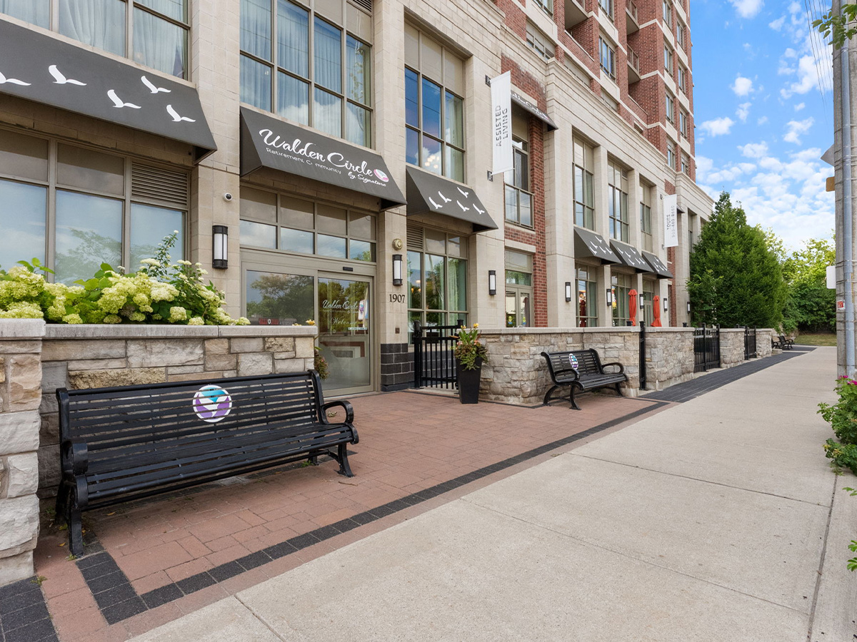 A bench in front of the exterior of Walden Circle Retirement Home, surrounded by greenery and a clear sky.